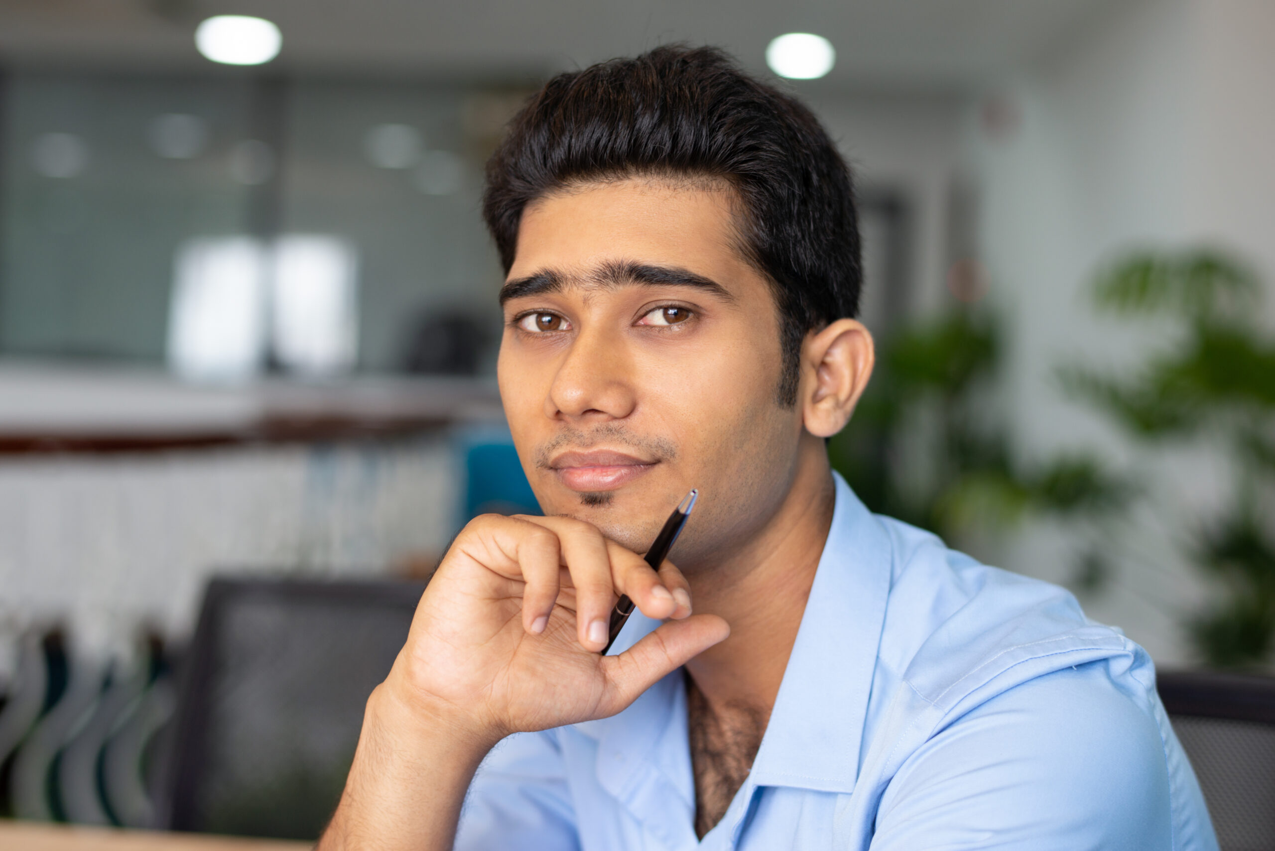 Portrait of young Indian businessman or student sitting with pen and looking at camera in office. Businessman concept